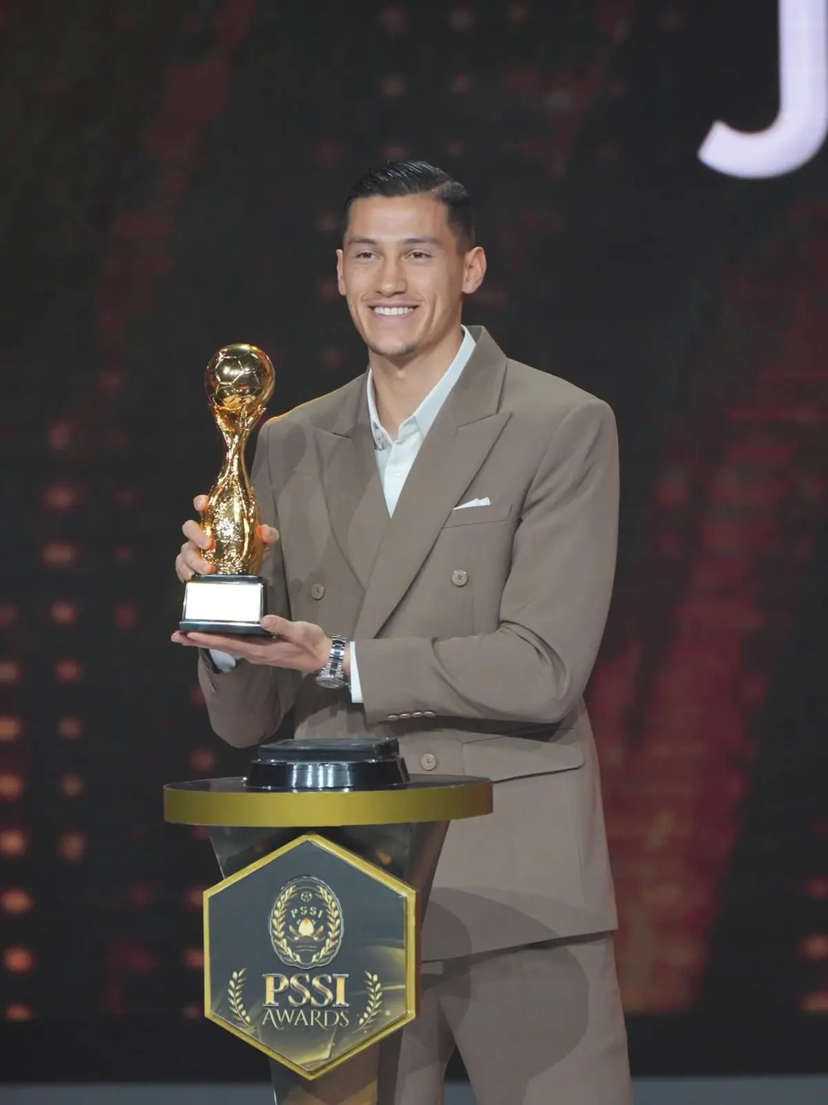 Jay Idzes mengenakan setelan jas berwarna cokelat muda sedang memegang trofi emas PSSI Awards 2026 dengan senyum bangga di atas panggung.