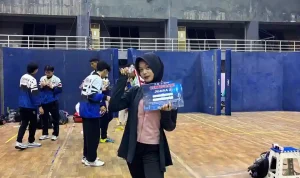 alya-rubiya-yuki-medali-perak-kejurnas-anggar-2026 Atlet anggar putri Alya Rubiya Yuki mengenakan hijab hitam dan jaket olahraga, berdiri di tengah lapangan GOR sambil memegang sertifikat Juara 2 dan menunjukkan medali peraknya.