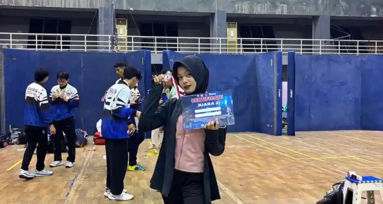 alya-rubiya-yuki-medali-perak-kejurnas-anggar-2026 Atlet anggar putri Alya Rubiya Yuki mengenakan hijab hitam dan jaket olahraga, berdiri di tengah lapangan GOR sambil memegang sertifikat Juara 2 dan menunjukkan medali peraknya.