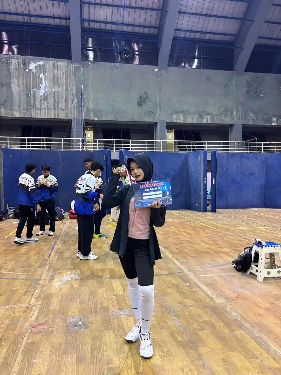 alya-rubiya-yuki-medali-perak-kejurnas-anggar-2026 Atlet anggar putri Alya Rubiya Yuki mengenakan hijab hitam dan jaket olahraga, berdiri di tengah lapangan GOR sambil memegang sertifikat Juara 2 dan menunjukkan medali peraknya.