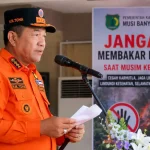 bupati-muba-toha-tohet-imbauan-larangan-membakar-lahan Bupati Musi Banyuasin H. M. Toha Tohet dengan seragam taktis BPBD berwarna oranye terang dan topi komando hitam-oranye bertuliskan H.M. TOHA sedang berbicara dengan tegas di depan mikrofon sambil memegang draf naskah pidato. Di latar belakang sebelah kanan, terdapat banner imbauan resmi dari Pemerintah Kabupaten Musi Banyuasin berwarna merah putih hitam mencolok bertuliskan JANGAN MEMBAKAR LAHAN! SAAT MUSIM KEMARAU disertai logo stop tangan dan visual ilustrasi kebakaran hutan.