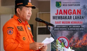 Bupati Musi Banyuasin H. M. Toha Tohet dengan seragam taktis BPBD berwarna oranye terang dan topi komando hitam-oranye bertuliskan H.M. TOHA sedang berbicara dengan tegas di depan mikrofon sambil memegang draf naskah pidato. Di latar belakang sebelah kanan, terdapat banner imbauan resmi dari Pemerintah Kabupaten Musi Banyuasin berwarna merah putih hitam mencolok bertuliskan JANGAN MEMBAKAR LAHAN! SAAT MUSIM KEMARAU disertai logo stop tangan dan visual ilustrasi kebakaran hutan.