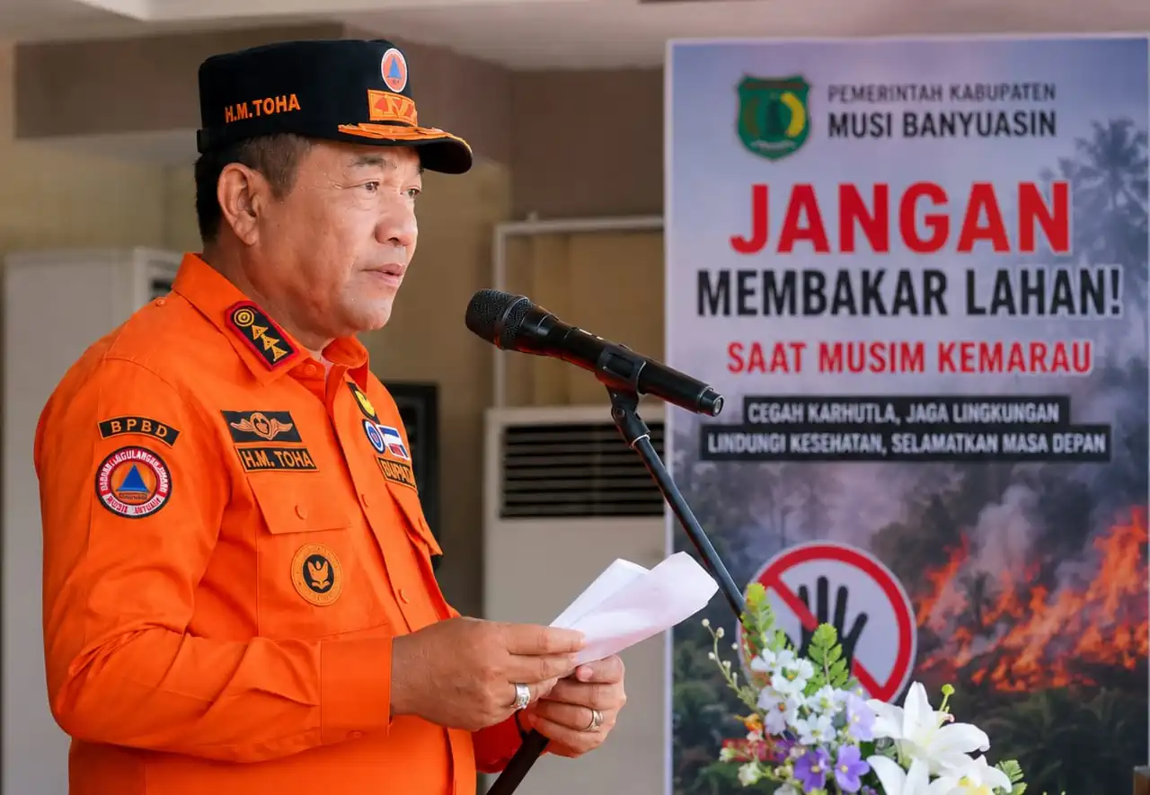 bupati-muba-toha-tohet-imbauan-larangan-membakar-lahan Bupati Musi Banyuasin H. M. Toha Tohet dengan seragam taktis BPBD berwarna oranye terang dan topi komando hitam-oranye bertuliskan H.M. TOHA sedang berbicara dengan tegas di depan mikrofon sambil memegang draf naskah pidato. Di latar belakang sebelah kanan, terdapat banner imbauan resmi dari Pemerintah Kabupaten Musi Banyuasin berwarna merah putih hitam mencolok bertuliskan JANGAN MEMBAKAR LAHAN! SAAT MUSIM KEMARAU disertai logo stop tangan dan visual ilustrasi kebakaran hutan.