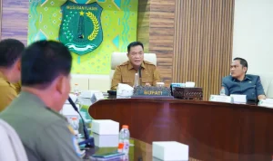 bupati-muba-toha-tohet-rapat-forkopimda-minyak-rakyat Bupati Muba H M Toha Tohet SH mengenakan seragam dinas cokelat aparatur sipil negara, duduk di kursi utama di tengah ruang rapat formal, berbicara di depan mikrofon meja. Di belakangnya terdapat logo tameng hijau besar Pemkab Musi Banyuasin dan jajaran peserta rapat menyimak dari sisi meja oval.