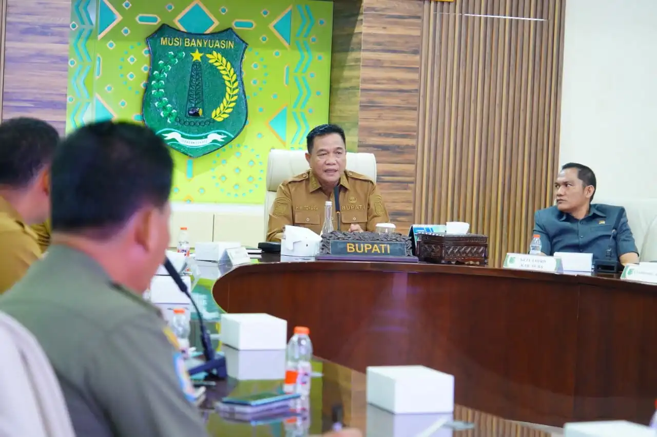 bupati-muba-toha-tohet-rapat-forkopimda-minyak-rakyat Bupati Muba H M Toha Tohet SH mengenakan seragam dinas cokelat aparatur sipil negara, duduk di kursi utama di tengah ruang rapat formal, berbicara di depan mikrofon meja. Di belakangnya terdapat logo tameng hijau besar Pemkab Musi Banyuasin dan jajaran peserta rapat menyimak dari sisi meja oval.