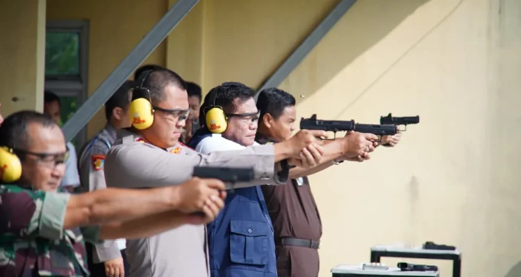Bupati Musi Banyuasin H. M. Toha Tohet, S.H. mengenakan rompi biru navy di atas kemeja putih, berkacamata pelindung, dan memakai earmuff kuning, sedang membidik sasaran menggunakan pistol dengan kedua tangan lurus ke depan di lapangan tembak terbuka. Di barisannya, tampak Kapolres Muba AKBP Ruri Prastowo dan jajaran Forkopimda lainnya juga sedang dalam posisi menembak dengan fokus yang sama.