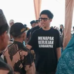 handry-pratama-putra-dprd-sumsel-ogan-ilir Anggota DPRD Provinsi Sumsel H. Handry Pratama Putra memakai kaos hitam bertuliskan Menapak Berjejak Berdampak sedang berbincang dengan warga di bawah tenda acara.