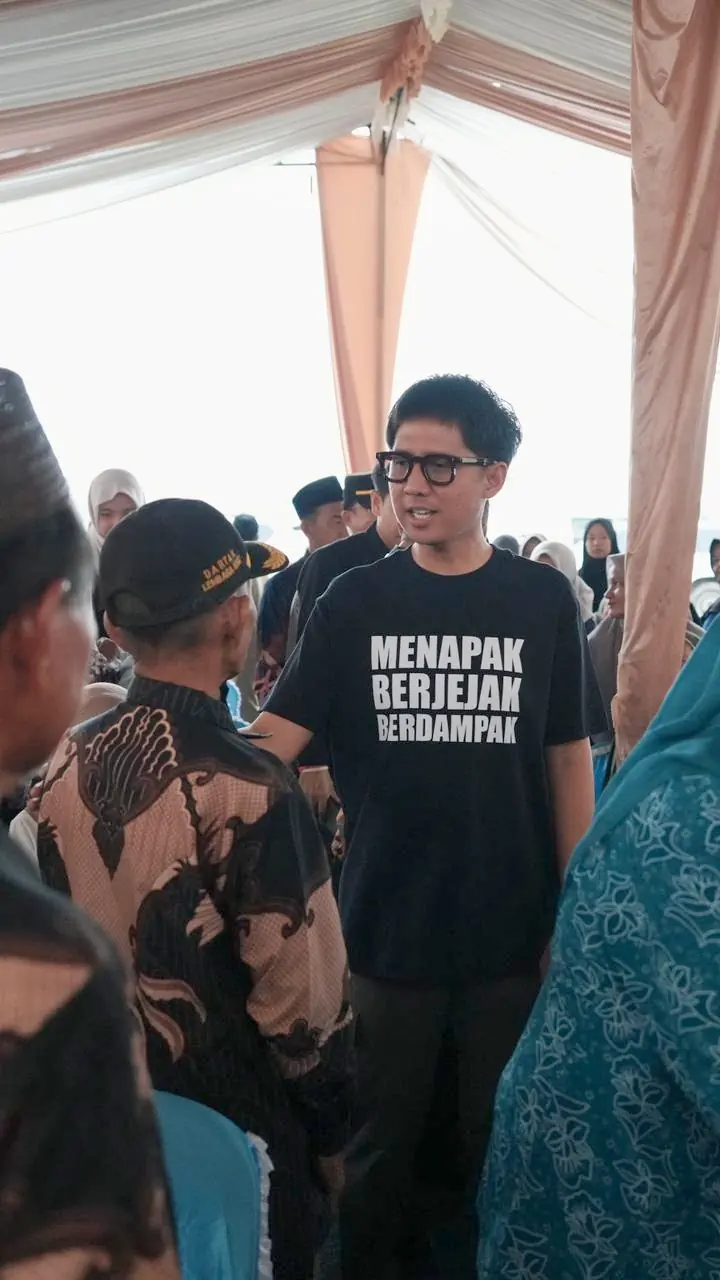 handry-pratama-putra-dprd-sumsel-ogan-ilir Anggota DPRD Provinsi Sumsel H. Handry Pratama Putra memakai kaos hitam bertuliskan Menapak Berjejak Berdampak sedang berbincang dengan warga di bawah tenda acara.