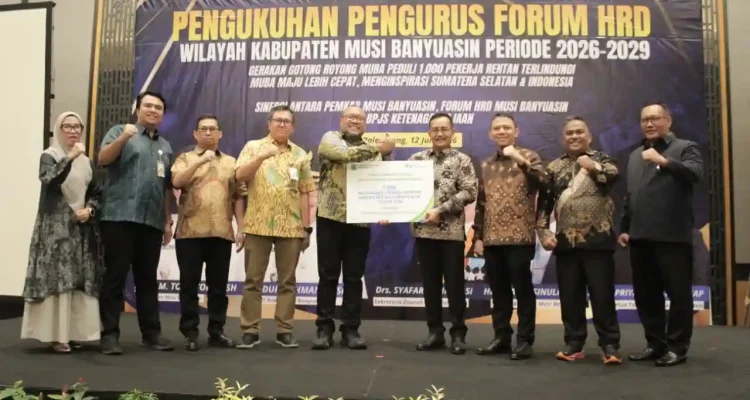 pelantikan-pengurus-forum-hrd-muba-2026 Jajaran pengurus Forum HRD Muba bersama pejabat pemkab dan direksi BPJS Ketenagakerjaan berdiri bersama di atas panggung formal, mengepalkan tangan ke depan sebagai simbol sinergi, sambil memegang maket kartu kepesertaan BPJS berukuran besar.