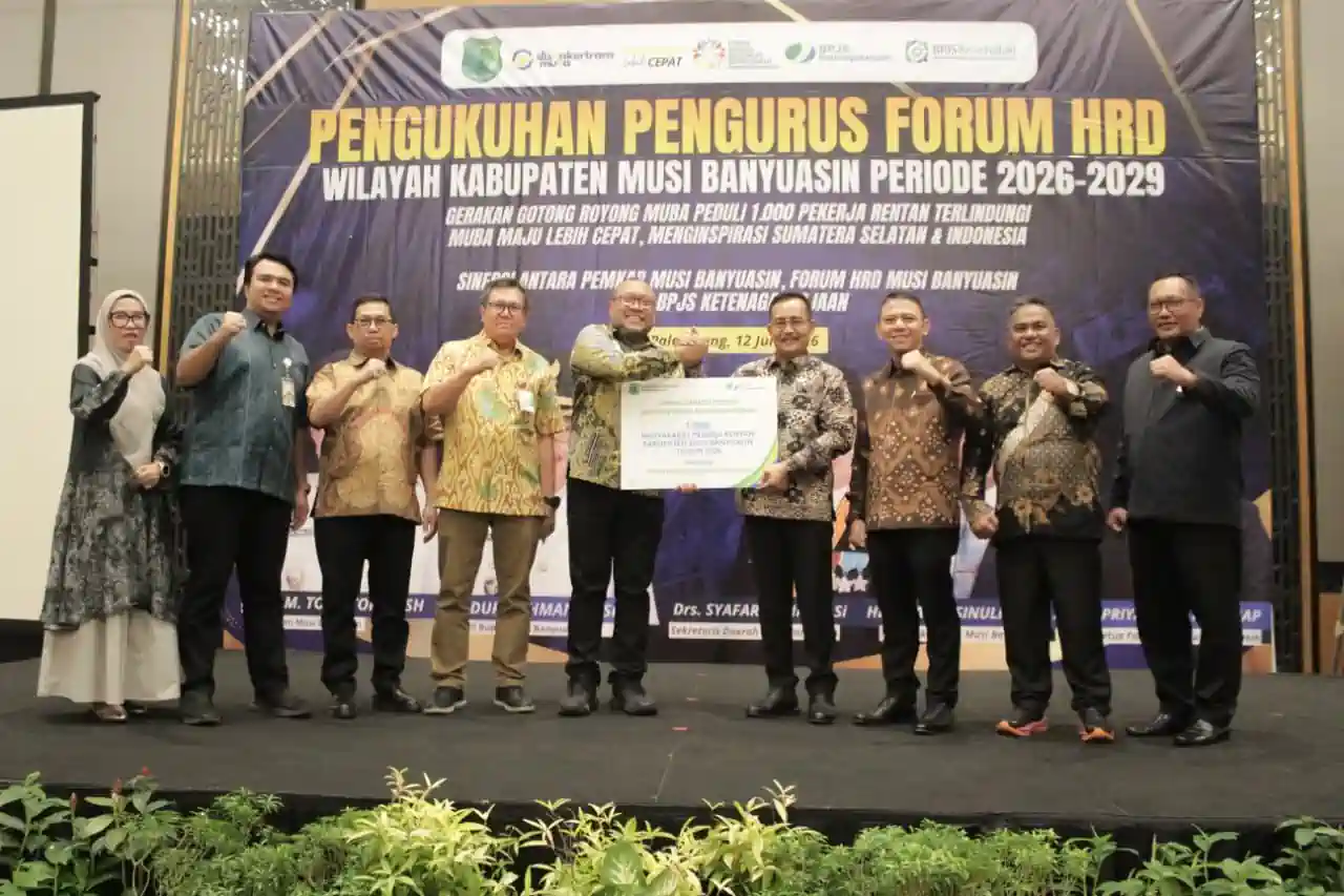 pelantikan-pengurus-forum-hrd-muba-2026 Jajaran pengurus Forum HRD Muba bersama pejabat pemkab dan direksi BPJS Ketenagakerjaan berdiri bersama di atas panggung formal, mengepalkan tangan ke depan sebagai simbol sinergi, sambil memegang maket kartu kepesertaan BPJS berukuran besar.