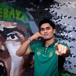 Penyerang baru Persebaya Surabaya Ramadhan Sananta berpose mengenakan jersey hijau sembari melakukan gestur telepon ikonik di depan mural suporter.
