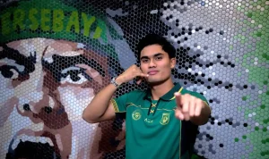 Penyerang baru Persebaya Surabaya Ramadhan Sananta berpose mengenakan jersey hijau sembari melakukan gestur telepon ikonik di depan mural suporter.