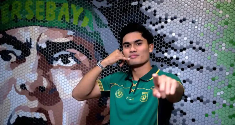 Penyerang baru Persebaya Surabaya Ramadhan Sananta berpose mengenakan jersey hijau sembari melakukan gestur telepon ikonik di depan mural suporter.