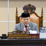 Wakil Ketua DPRD Provinsi Sumatera Selatan Ilyas Panji Alam mengenakan jas abu-abu dan peci hitam sedang memimpin jalannya Rapat Paripurna dari atas podium utama yang berornamen ukiran kayu khas Palembang.