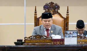 rapat-paripurna-dprd-sumsel-apbd-2025 Wakil Ketua DPRD Provinsi Sumatera Selatan Ilyas Panji Alam mengenakan jas abu-abu dan peci hitam sedang memimpin jalannya Rapat Paripurna dari atas podium utama yang berornamen ukiran kayu khas Palembang.