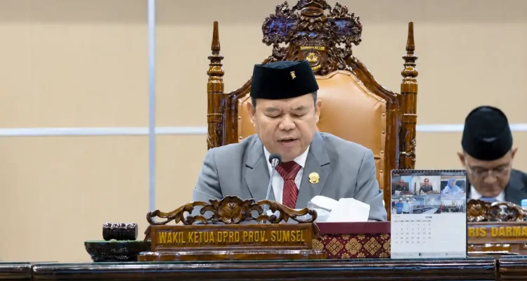 rapat-paripurna-dprd-sumsel-apbd-2025 Wakil Ketua DPRD Provinsi Sumatera Selatan Ilyas Panji Alam mengenakan jas abu-abu dan peci hitam sedang memimpin jalannya Rapat Paripurna dari atas podium utama yang berornamen ukiran kayu khas Palembang.