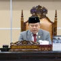 Wakil Ketua DPRD Provinsi Sumatera Selatan Ilyas Panji Alam mengenakan jas abu-abu dan peci hitam sedang memimpin jalannya Rapat Paripurna dari atas podium utama yang berornamen ukiran kayu khas Palembang.