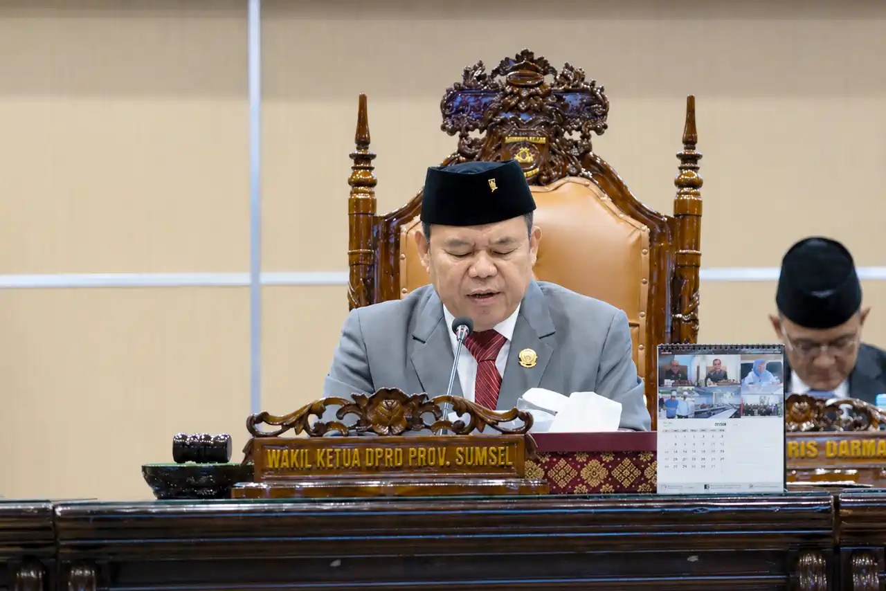 rapat-paripurna-dprd-sumsel-apbd-2025 Wakil Ketua DPRD Provinsi Sumatera Selatan Ilyas Panji Alam mengenakan jas abu-abu dan peci hitam sedang memimpin jalannya Rapat Paripurna dari atas podium utama yang berornamen ukiran kayu khas Palembang.