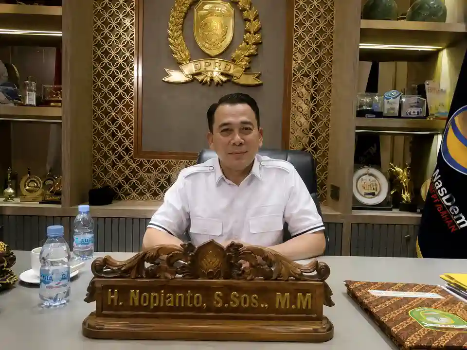 Wakil Ketua DPRD Sumsel H Nopianto duduk di ruang kerjanya dengan latar belakang lambang DPRD dan bendera Partai NasDem.